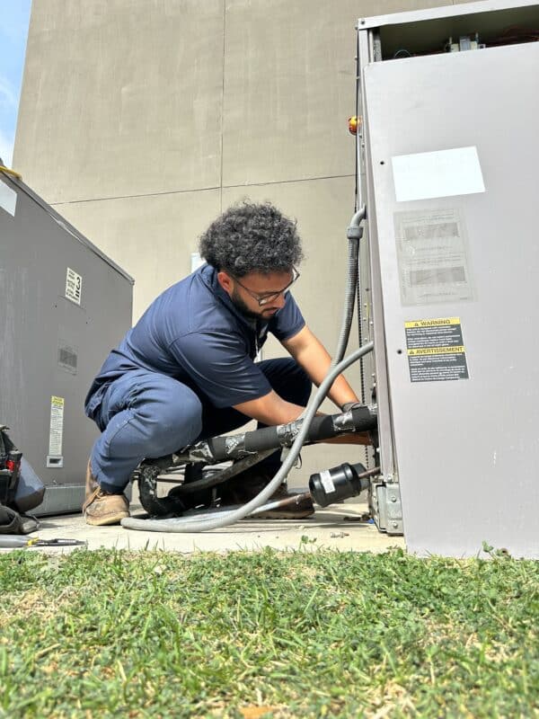 A man wearing a navy blue work outfits squatting down at the base of an exterior HVAC unit performing work.