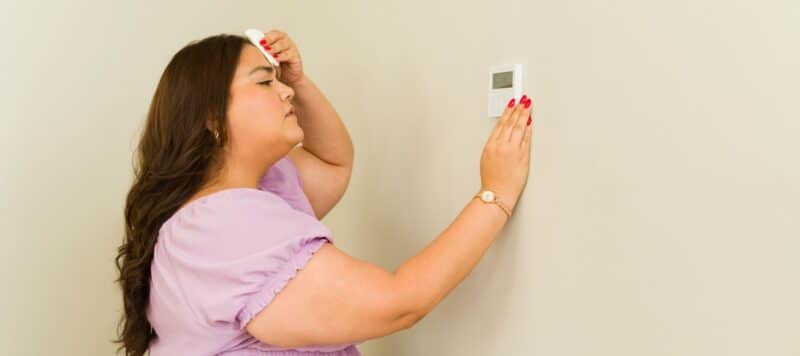 woman holding her hand up to her head in discomfort while her other hand is adjusting a digital thermostat